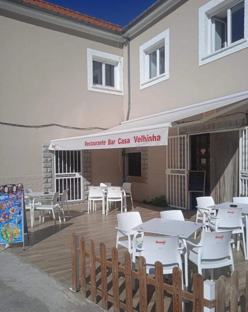 Esplanada com mesas brancas e toldo, entrada do Restaurante Bar Casa Velhinha, edifício bege com janelas brancas