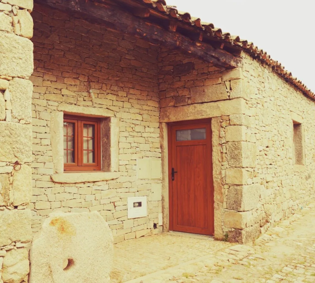 Casa tradicional de pedra clara com porta e janela vermelhas, telhado em telha cerâmica, piso de calçada irregular.