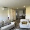 Sala de estar moderna com dois sofás brancos, tapete padrão em cinzentos e vermelhos, e cozinha aberta ao fundo com mesa de jantar.