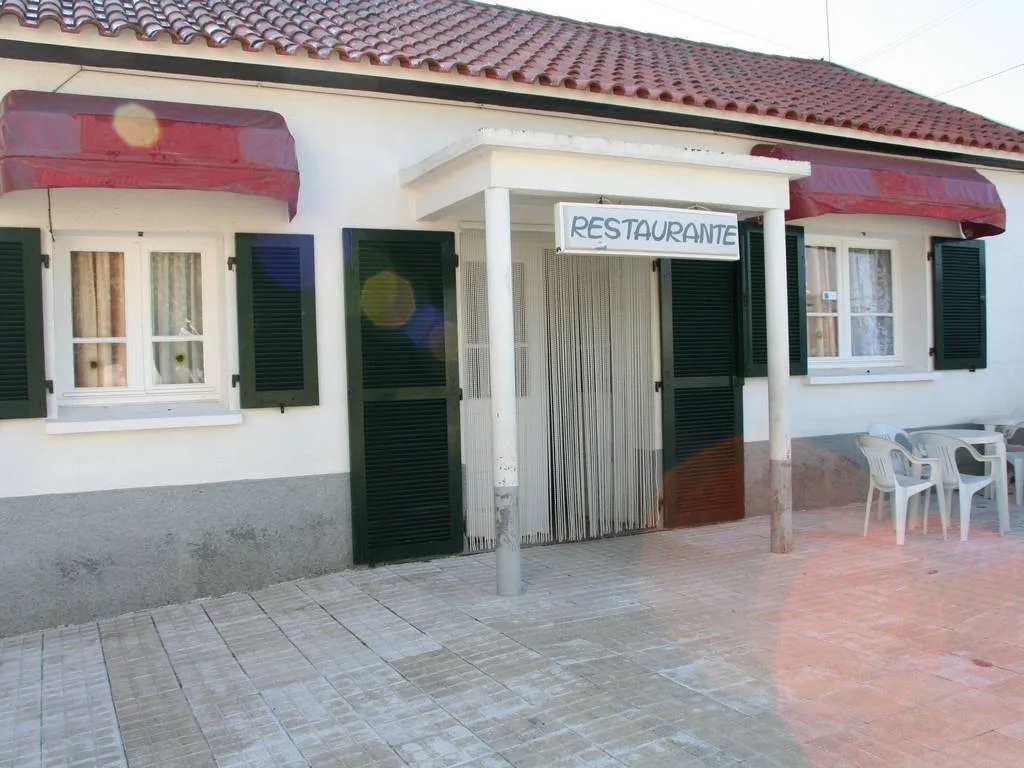 Fachada de restaurante com entrada central, janelas com venezianas verdes, toldo vermelho e esplanada lateral com cadeiras brancas.