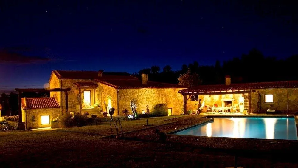 Casa de campo portuguesa iluminada à noite, com piscina refletindo as luzes, rodeada de paisagem rural escura.